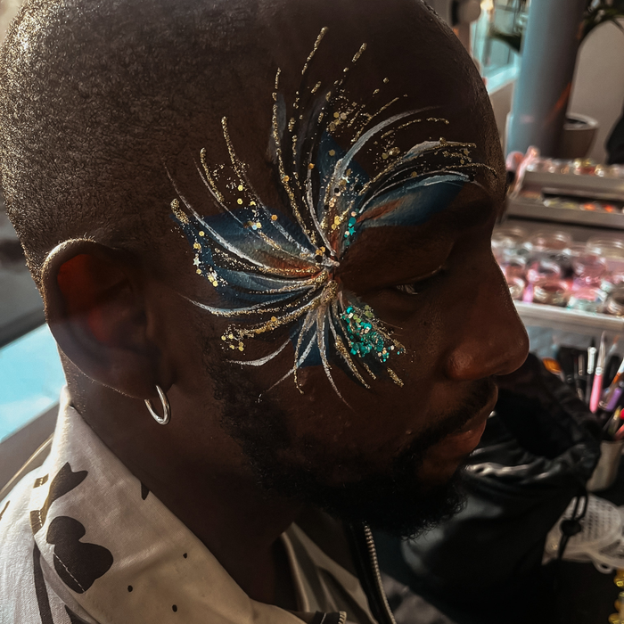 A close-up side profile of a man with elaborate face paint featuring a blue, gold, and teal butterfly or wing-like design on his forehead, in a makeup studio setting.
