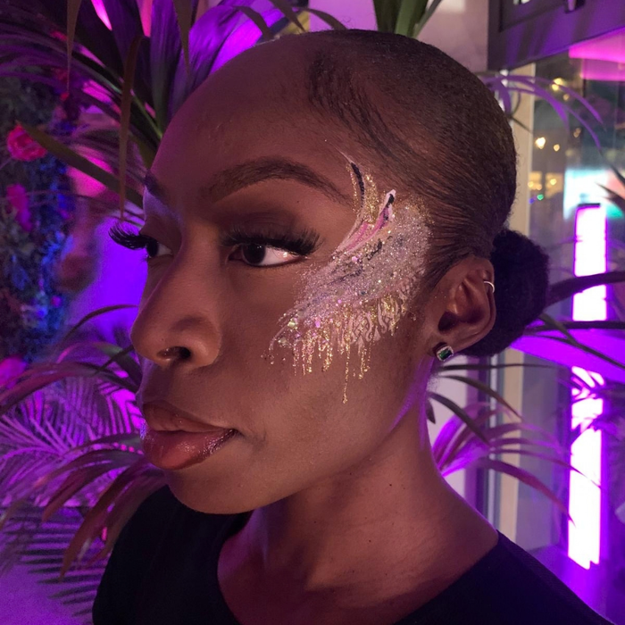 A close-up portrait of a woman with dramatic glittery gold and silver face paint resembling a feather, styled with a high bun, set against a background of purple ambient lighting and tropical plants.