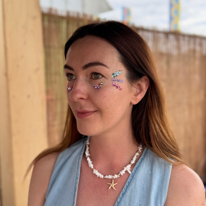 A woman with brown hair is shown in profile, wearing a light blue sleeveless top, a white shell necklace with a gold starfish pendant, and colorful glittery eye stickers. The background features a wooden fence and a light-colored wall.