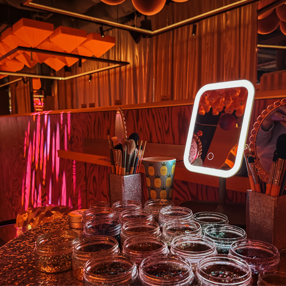 A vanity-style makeup station with a lit square mirror, makeup brushes in holders, and multiple small clear jars containing loose glitter or powder, set in a wood-paneled room with warm ambient lighting.