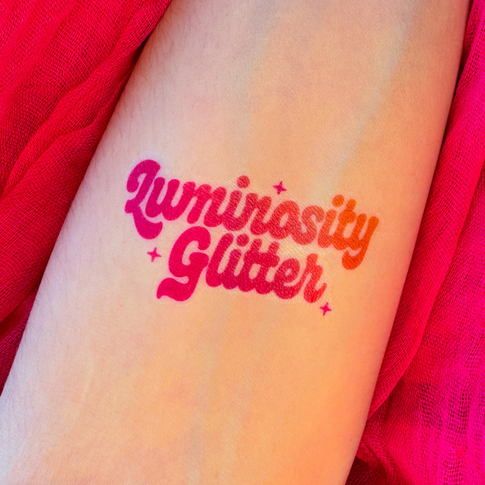 A temporary tattoo on a person's forearm reading 'Luminosity Glitter' in a pink-to-orange gradient script, surrounded by bright pink fabric.