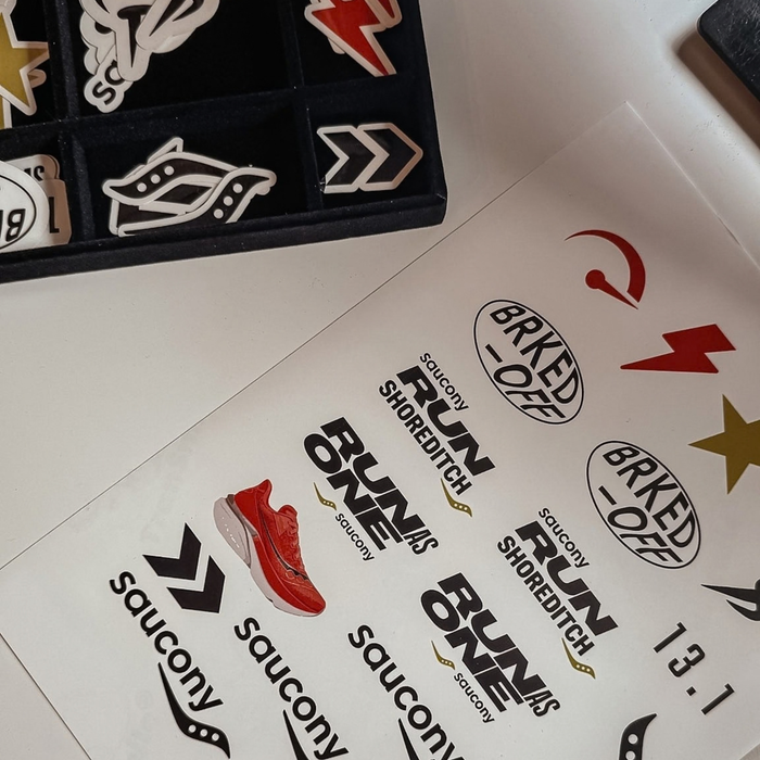 A close-up of a sheet of Saucony branded decals or stickers laid on a white surface, with a black tray of additional decals partially visible at the top left. The sheet includes multiple logo variations, including 'Saucony', 'Run Shoreditch', 'Run One', 'Barked Off', and '13.
