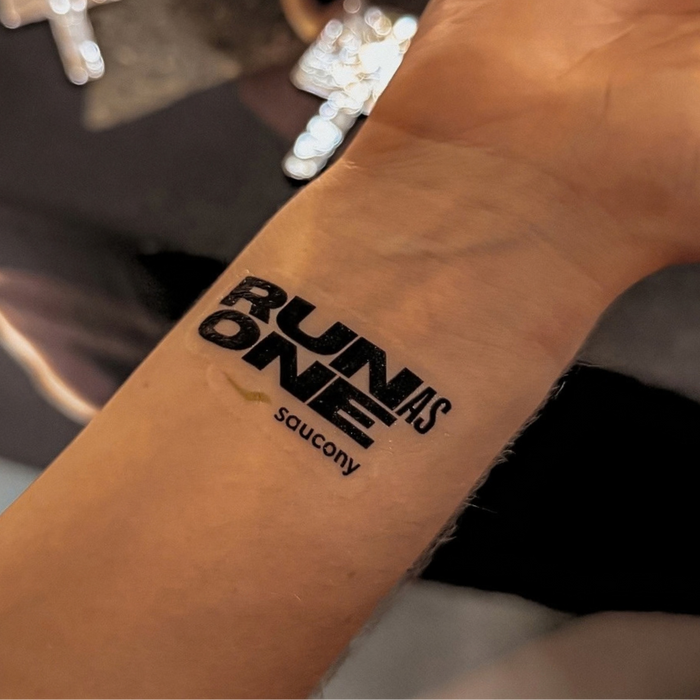 A close-up of a person's inner forearm with a black temporary tattoo reading 'RUN AS ONE' and 'saucony' underneath, showing the skin tone and part of a dark shirt in the background.