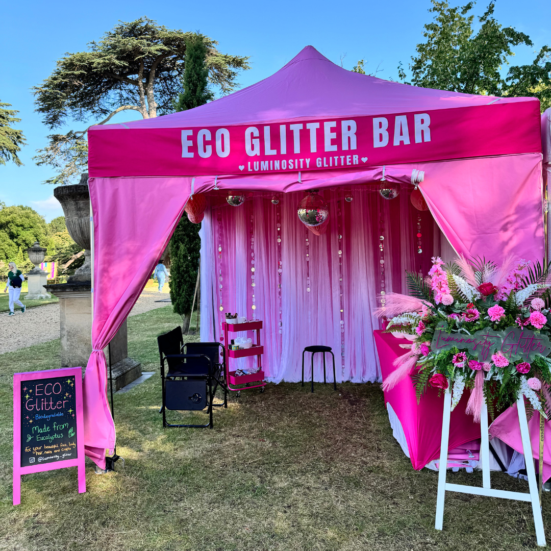 A pink pop-up canopy tent set up outdoors on grass, decorated as an event booth with white lettering that reads 'ECO GLITTER BAR' and 'LUMINOSITY GLITTER'. The canopy is draped in pink fabric, with a pink-to-white gradient curtain backdrop inside, small chairs, a mini cart, and floral arrangements in the foreground.