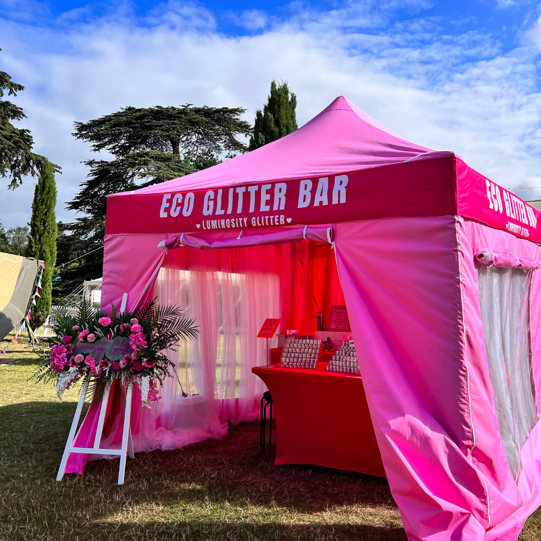 A bright pink event canopy tent with white lettering reading 'ECO GLITTER BAR' and 'LUMINITY GLITTER' set up outdoors on grass under a blue sky. A red display table sits inside the tent, and a floral arrangement stands on a white easel beside it.