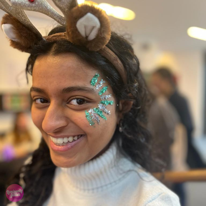 A smiling woman wearing a festive reindeer antler headband with brown faux fur and silver glitter trim, paired with green and silver glitter face paint on her cheek. She is wearing a white turtleneck sweater in a softly blurred indoor setting.