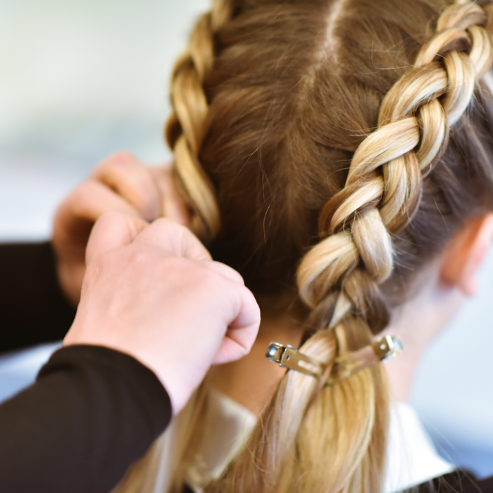 A close-up of a person's blonde hair styled in a braided ponytail with a hand adjusting it, showing the back of the head and hair details.