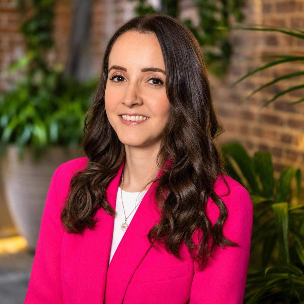 A woman with long, wavy brown hair is shown from the chest up, wearing a bright pink blazer over a white top. She is smiling and looking slightly off-camera.