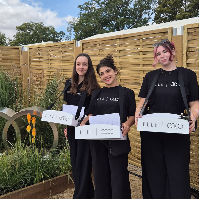Three women standing outdoors in front of a wooden slat fence, each holding a white box printed with the brand name 'ELLE' and the Audi four-ring logo. They are dressed in matching black outfits.