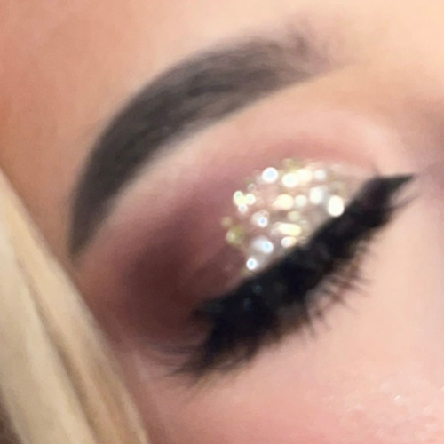 A close-up of a person's eye showcasing a glittery eye makeup look with a large, sparkling clear rhinestone or crystal embellishment on the upper lid, paired with black eyeliner and mascara.