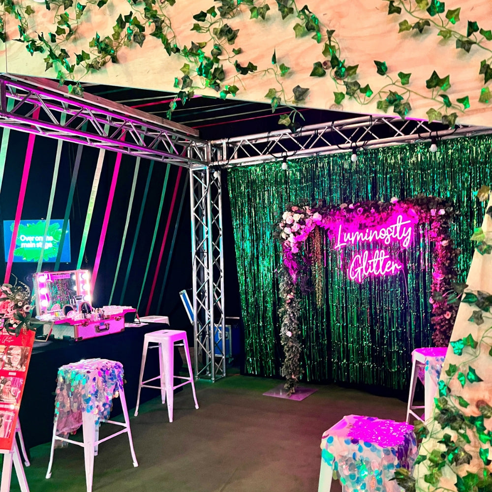 Decorative setup with greenery, neon lights, and a 'Luminosity Glitter' sign.