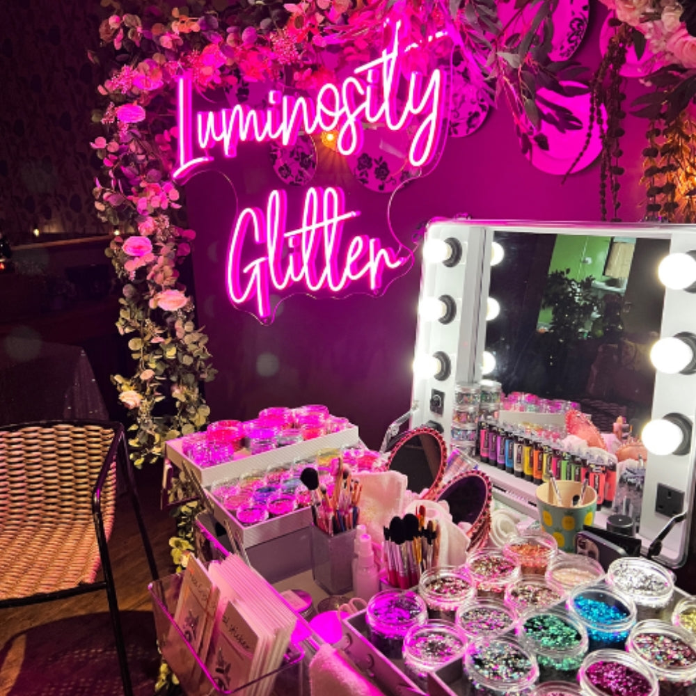 A decorated vanity setup with a large pink neon sign reading 'Luminosity Glitter' above a lighted makeup mirror, surrounded by jars of face gems, glitter, brushes, and cosmetics in a pink-lit room.