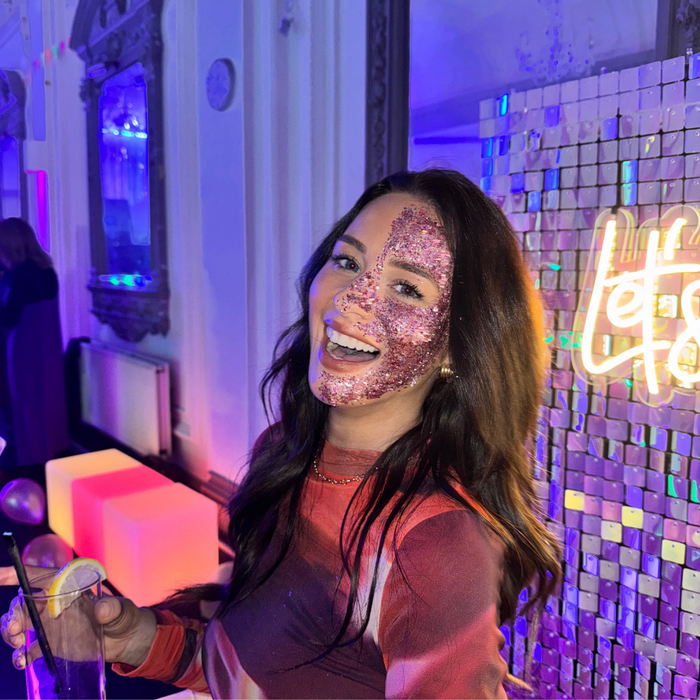A woman with a glittery pink face mask smiles in a brightly lit indoor venue with purple and blue ambient lighting, a mosaic wall, and a neon sign in the background.