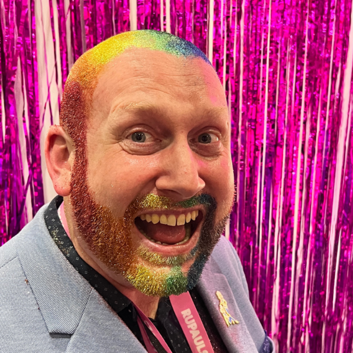 A smiling man with a rainbow-colored shaved head and beard stands in front of a bright pink tinsel backdrop, wearing a light blue blazer and a pink lanyard.