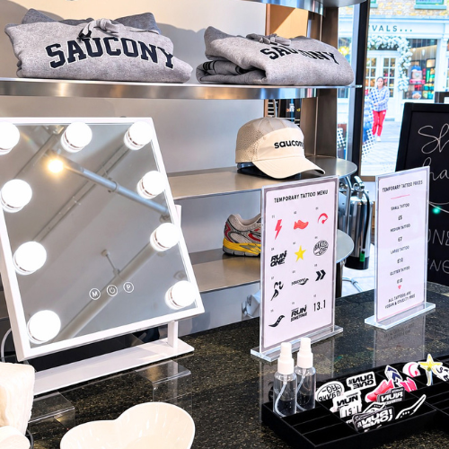 A retail display featuring a white vanity mirror with built-in light bulbs, small bottles, temporary tattoo stickers, and branded Saucony apparel including folded gray sweatshirts and a beige cap on shelves.