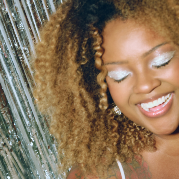 A close-up lifestyle shot of a smiling woman with voluminous curly brown hair, wearing silver glitter eyeshadow and a white top, standing in front of a silver tinsel curtain backdrop.