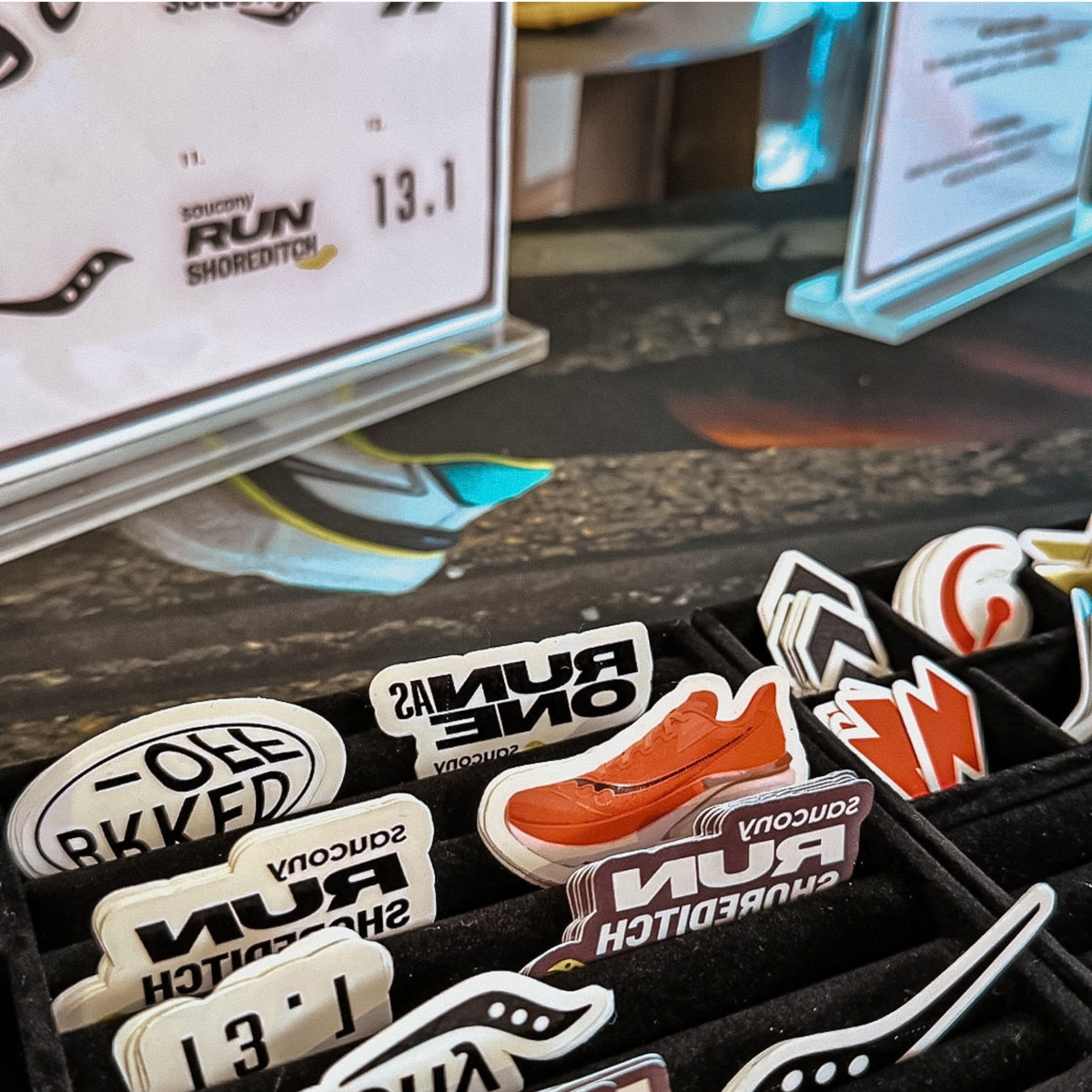 A close-up of a display of custom temporary tattoos featuring 'Run Shoreditch' and 'Saucony' designs, with a Saucony shoe pin in the center and a sign in the background.