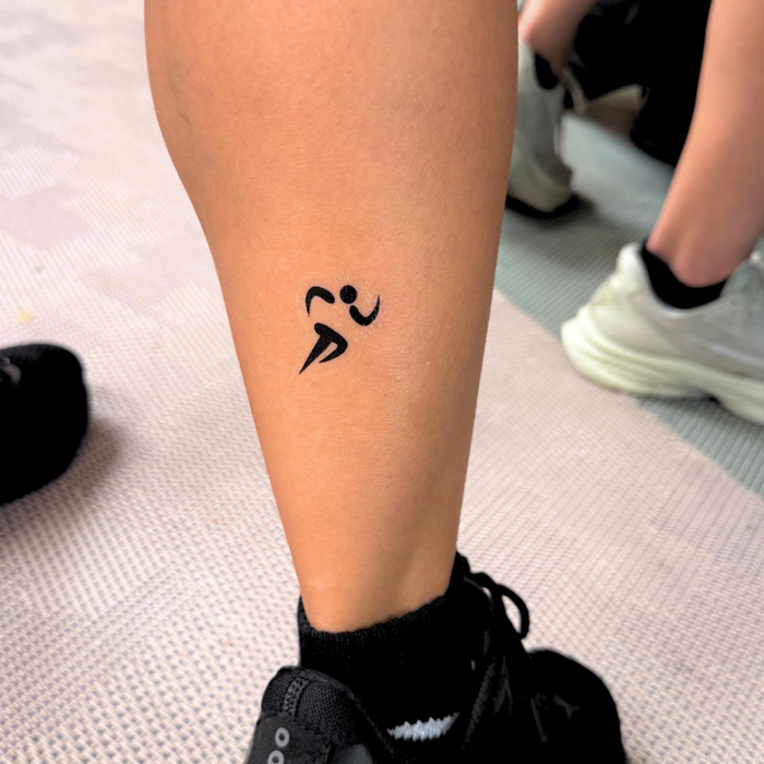A close-up of a person's calf with a small black minimalist running figure temporary tattoo, wearing black socks and black sneakers on a light textured floor.