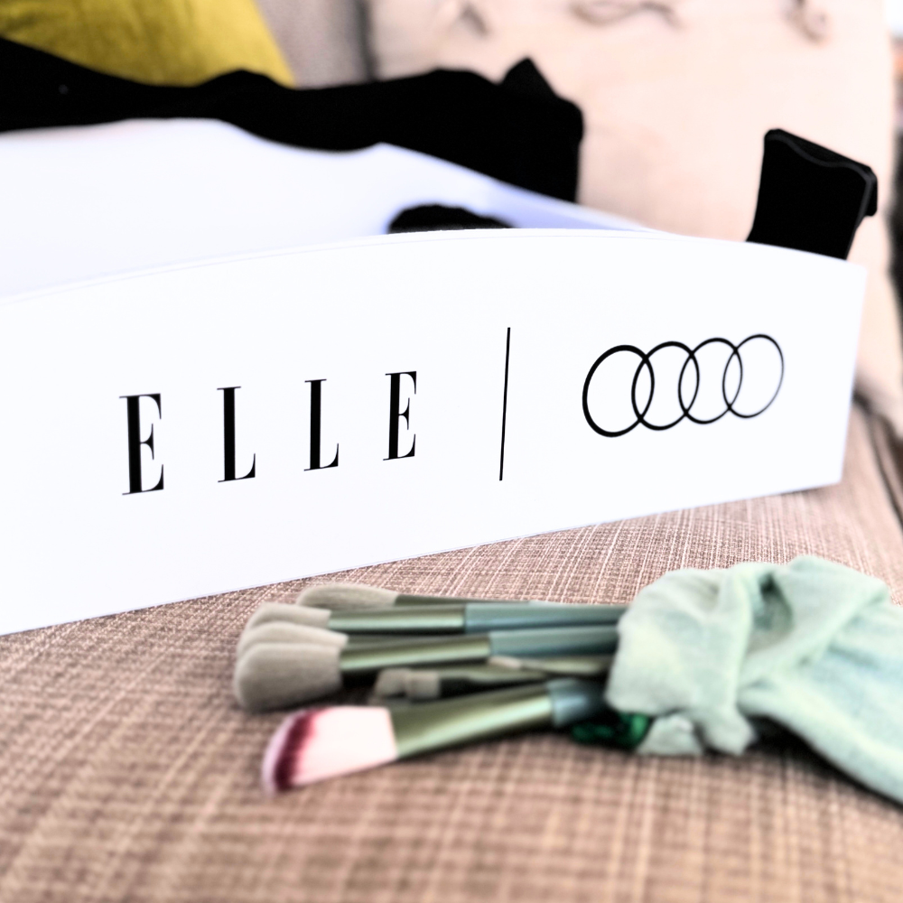 A white rectangular box with the brand name 'ELLE' and four interlocking rings logo printed in black, placed on a textured beige surface. Several makeup brushes with green handles and a pale mint green cloth are arranged in front of the box.